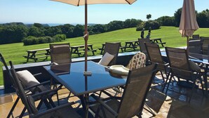 An outdoor patio with a table and chairs, a large umbrella, and a view of a grassy area. - Greenkeepers Cottage - UK6712 (Lydstep, near Tenby)