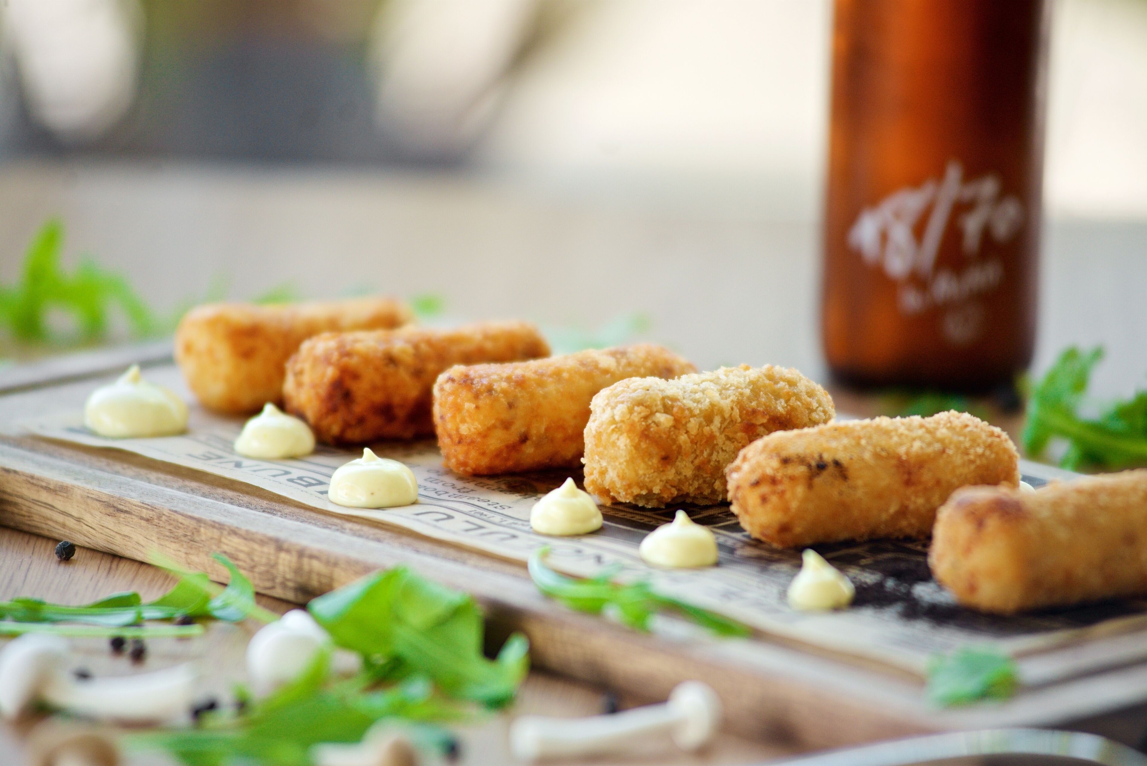 A wooden board with several breaded items and small dollops of sauce, accompanied by fresh greens and mushrooms.