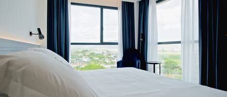 A hotel room with a large bed, a chair, and a small table. There are two windows with curtains, and a view of a cityscape is visible through the windows.