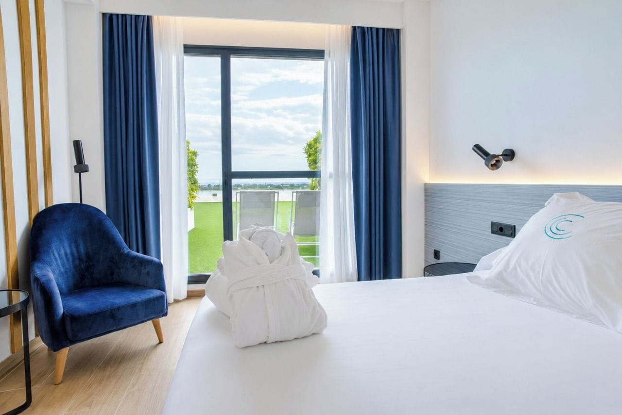 Habitación de hotel con una cama, una silla azul, una bata blanca y vistas al exterior.
