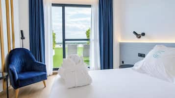 Habitación de hotel con una cama, una silla azul, una bata blanca y vistas al exterior.