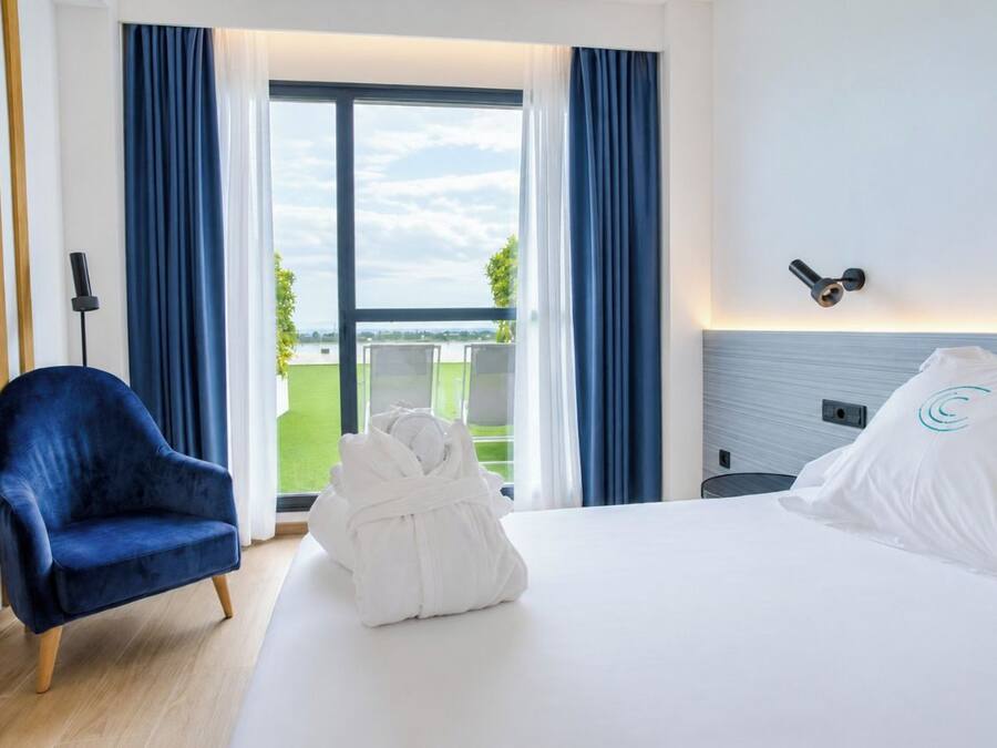 Habitación de hotel con una cama, una silla azul, una bata blanca y vistas al exterior.