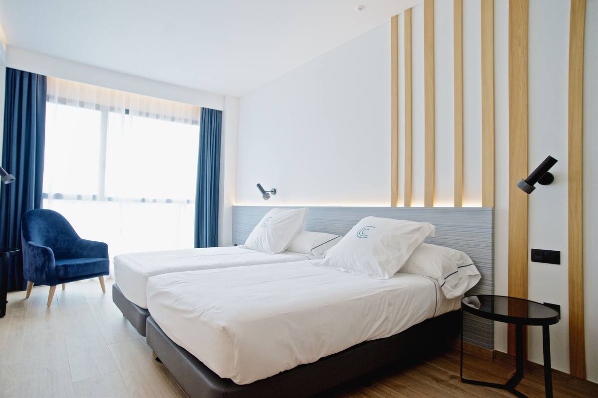 Une chambre d’hôtel avec deux lits, une chaise, une petite table et une fenêtre avec des rideaux.