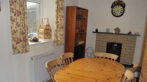 A dining room with a round wooden table surrounded by six chairs, a bookshelf, a window with floral curtains, and a fireplace with a clock above it. - 3 bedroom accommodation in Silverdale near Grange Over Sands (Silverdale near Grange Over Sands)