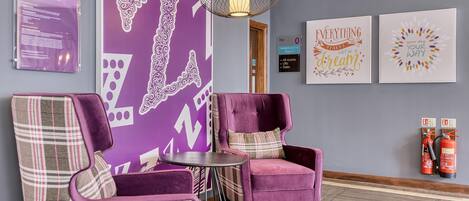 A lobby with purple chairs, a round table, a purple wall with white patterns, and a hanging light fixture.