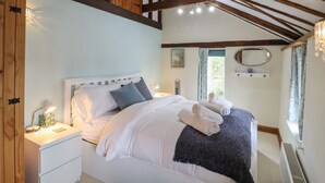 A bedroom with a large bed, wooden beams, a bedside table with a lamp, and a window with curtains. - The Old Custom House (Halesworth)