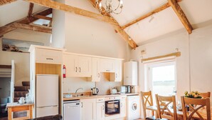 A kitchen with white cabinets, a refrigerator, a stove, and a microwave. There is a dining table with chairs and a chandelier. - Goose Cottage- UK6737 (Lydstep, near Tenby)