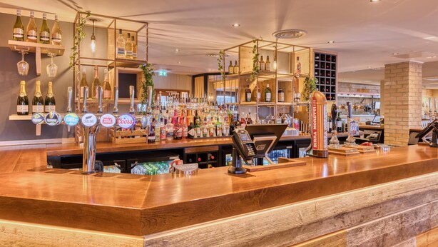 A bar area with a wooden counter, multiple liquor bottles, and a cash register. - Premier Inn Worksop (Worksop)