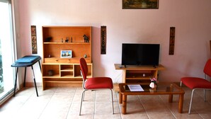 A living room with a TV, two red chairs, a wooden shelf with decorative items, and a small table with a vase. - Independent 25m2 studio in Villa: Wi-fi + terrace (Saint-Gély-du-Fesc)