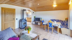 Un salon chaleureux avec un canapé, une table basse, une cheminée, un coin repas avec une table et des chaises, et une cuisine visible à travers une arche.