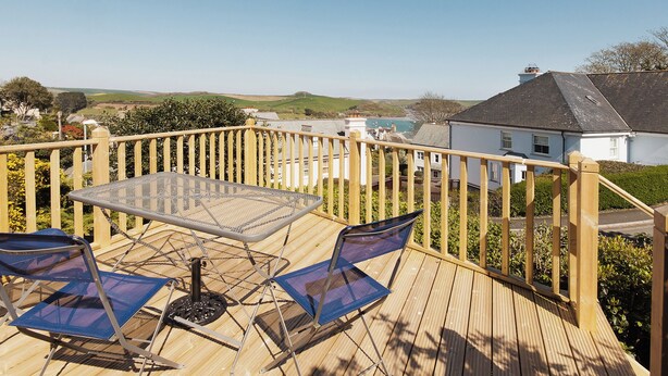 Terraza de madera con mesa de vidrio y dos sillas, con vista a una zona costera con casas y colinas.