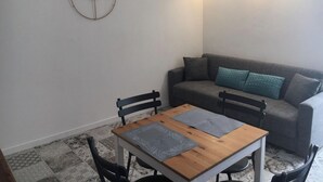 Un espacio de sala pequeño y moderno con una mesa de madera, dos sillas, un sofá gris y un cojín azul verdoso, y un reloj de pared.