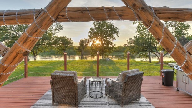 A wooden deck with wicker chairs and a table, overlooking a lake and trees.