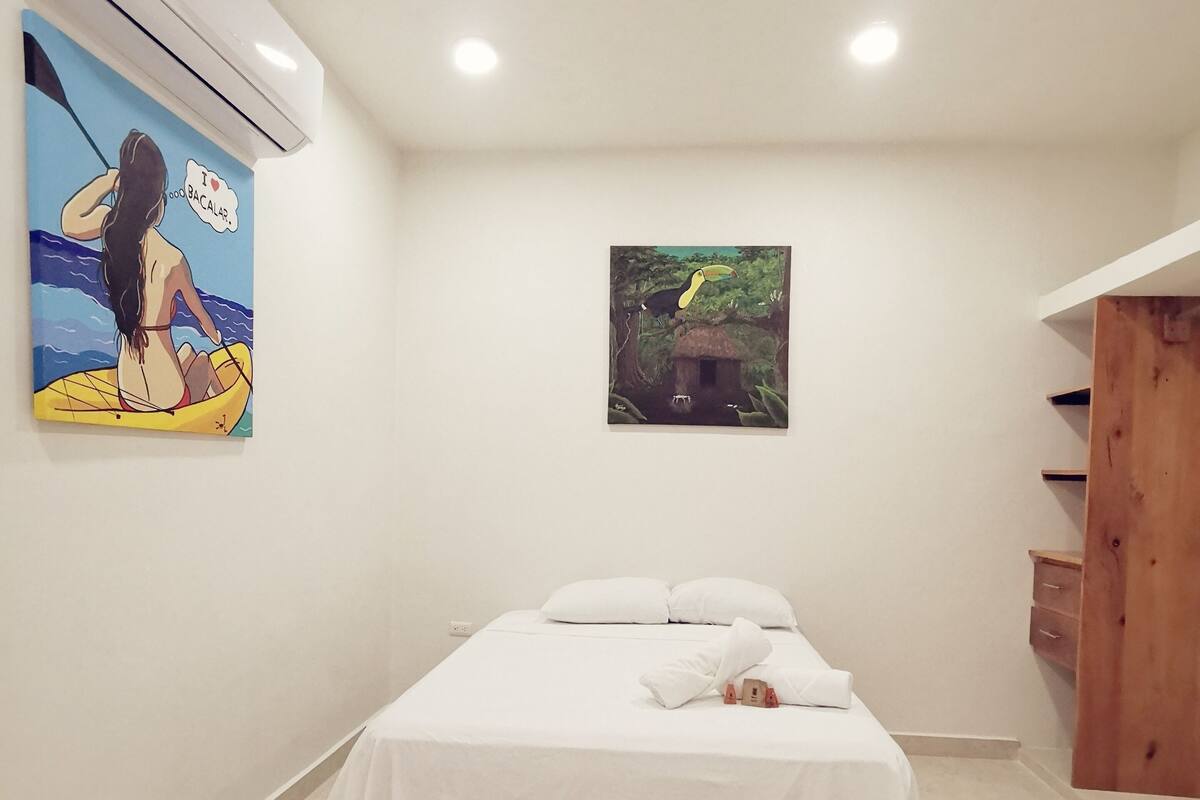 A bedroom with a bed, a painting of a woman on a boat, and a painting of a toucan.