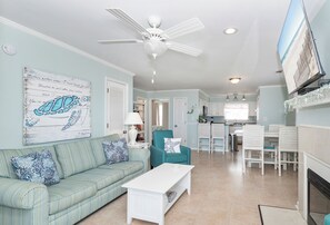 A living room with a ceiling fan, a sofa with patterned cushions, a white coffee table, and a kitchen area in the background.