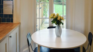 A kitchen with a white table and chairs, a vase with yellow flowers, and a window with a view of greenery. - 5 bedroom accommodation in Victoria Road (Victoria Road)
