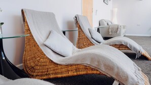 A room with two wicker lounge chairs, a glass table, a white chair, and a wall-mounted light fixture.