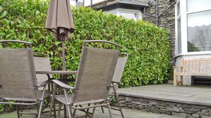 Une terrasse extérieure avec des chaises et une table en osier, un mur de pierre et une haie en arrière-plan.