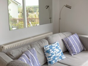 A living room with a grey sofa, two striped pillows, a radiator, and a window overlooking a balcony with plants and a chair. - 3 bedroom accommodation in Salcombe (Salcombe)