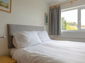 Un lit bien fait, avec du linge de lit blanc, dans une pièce dotée d’une tête de lit grise, d’une fenêtre avec des rideaux et d’un radiateur.