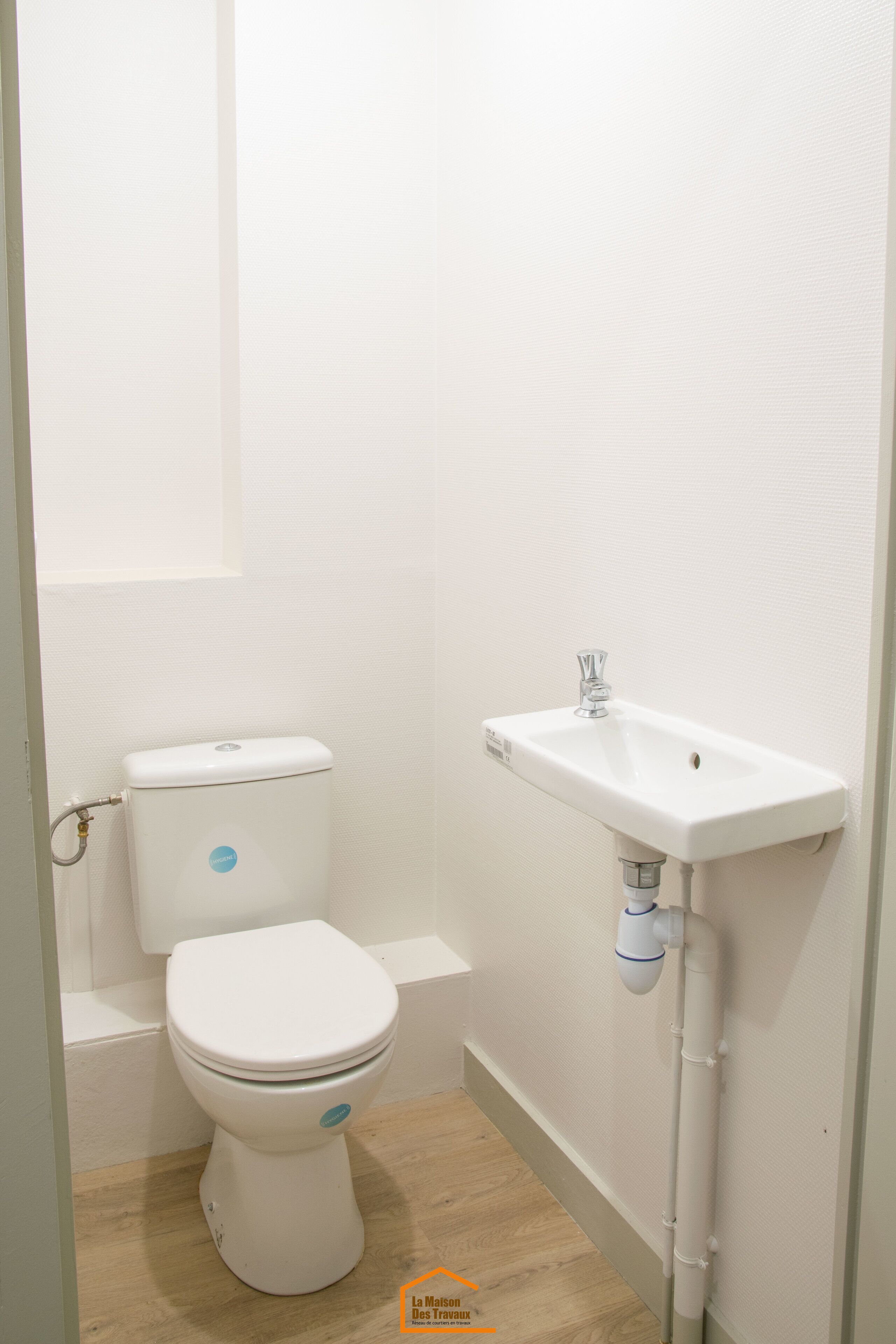 A small, clean bathroom with a white toilet and sink, wooden flooring, and a plain wall.