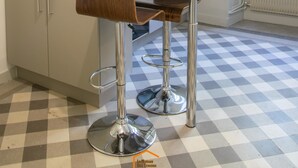A modern kitchen with a checkered floor, a wooden table, two bar stools, and white cabinetry. - Apartment du Puits Salé (Lons-le-Saunier)