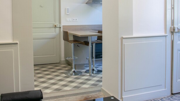A compact living space with a sofa, a glass coffee table, and a remote control on it. There is a checkered floor pattern and a door leading to a kitchen area. - Apartment du Puits Salé (Lons-le-Saunier)