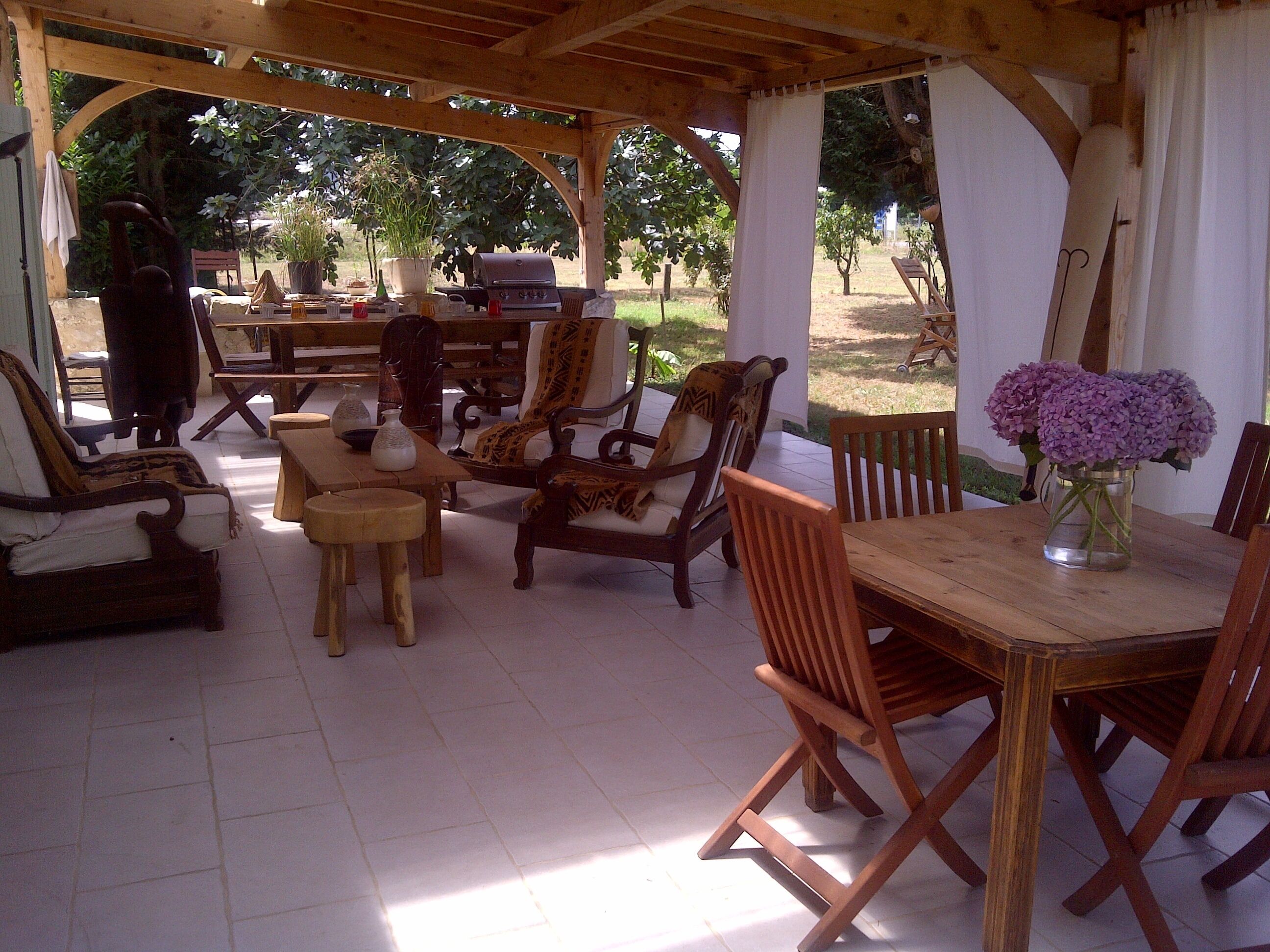 A covered outdoor seating area with wooden furniture, a grill, and a table with a vase of flowers.