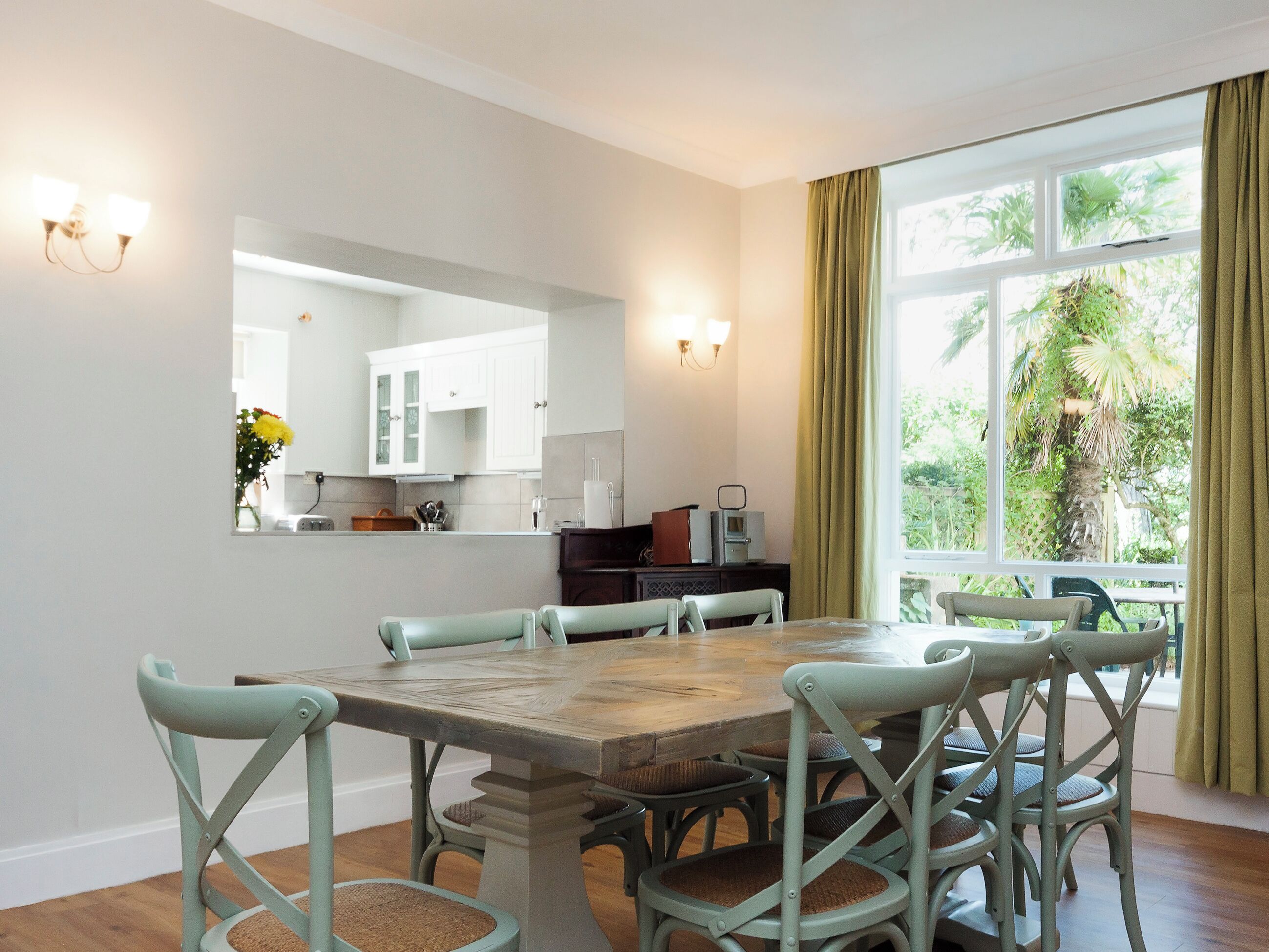 Un comedor con una mesa y sillas de madera, una cocina al fondo y un ventanal con cortinas.
