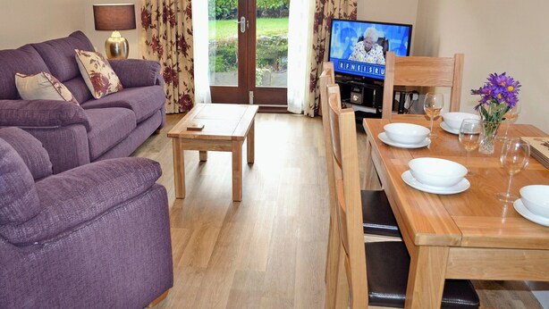 A living room with a purple sofa, a wooden coffee table, a TV, and a dining area with a wooden table set for two. - 2 bedroom accommodation in Bowness on Windermere (Bowness on Windermere)