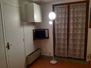 Huoneessa on valkoinen vaatekaappi, pieni seinälle asennettu televisio, lattiavalaisin ja kuviollinen verho.