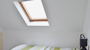 Una habitación con tragaluz, una cama con colcha a rayas, una mesita de noche con lámpara y una mesita auxiliar de madera.