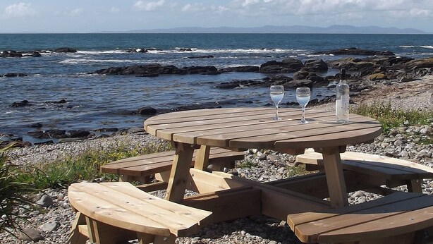 Mesa de picnic de madera con dos vasos y una botella, ubicada en una playa rocosa con el océano y montañas al fondo.