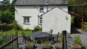 A white building with a grey roof, a small porch with a red railing, and a round table with chairs in front. - 3 bedroom accommodation in Satterthwaite (Satterthwaite)