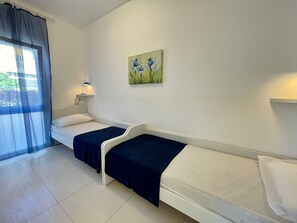A room with two beds, a window with sheer curtains, a wall-mounted lamp, and a painting of flowers.