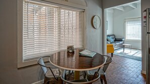 A dining area with a round table, chairs, a clock on the wall, and a window with blinds.