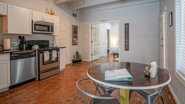 A kitchen with white cabinets, stainless steel appliances, a round dining table, and a brick floor.