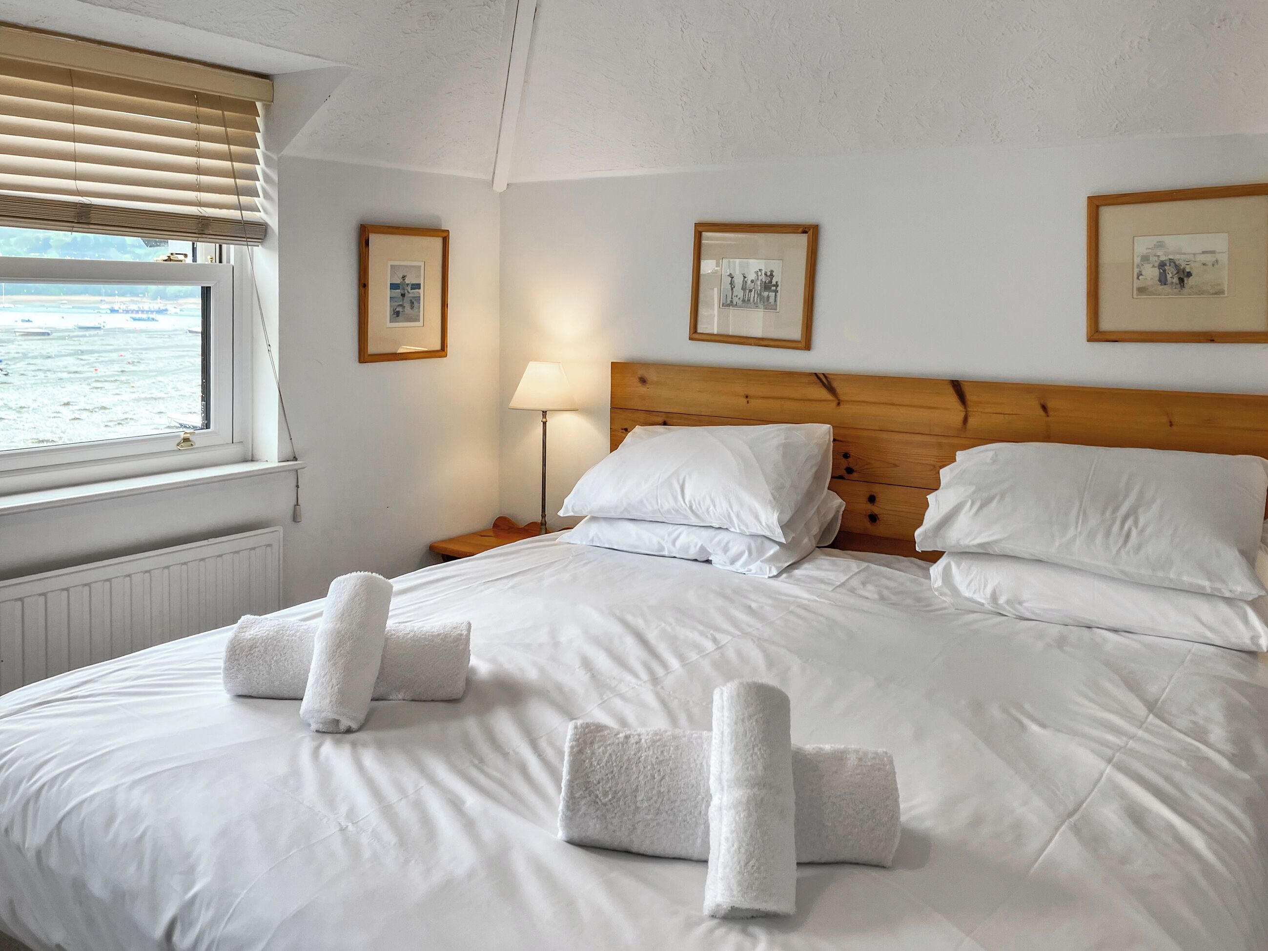 A neatly made bed with white linens and towels, a wooden headboard, a bedside lamp, and framed pictures on the wall.