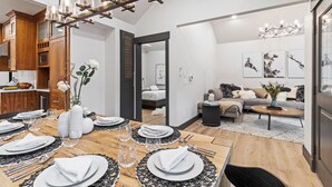 Un comedor moderno con una mesa de madera dispuesta para una comida, una araña colgante y vistas a un dormitorio con una cama y un sofá.