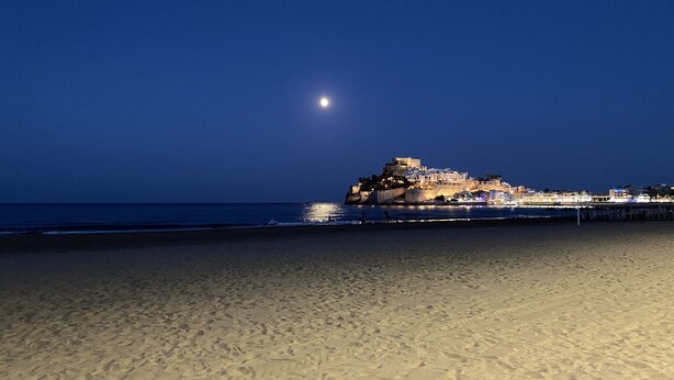A beach with a historic building on a hill, a pier extending into the water, and a moon in the sky.