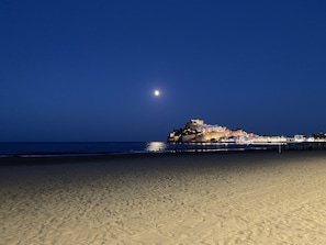 A beach with a historic building on a hill, a pier extending into the water, and a moon in the sky.