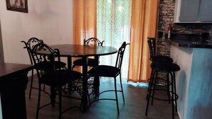 Un coin repas avec une table ronde et six chaises, un mur en pierre et une fenêtre ornée de rideaux orange.