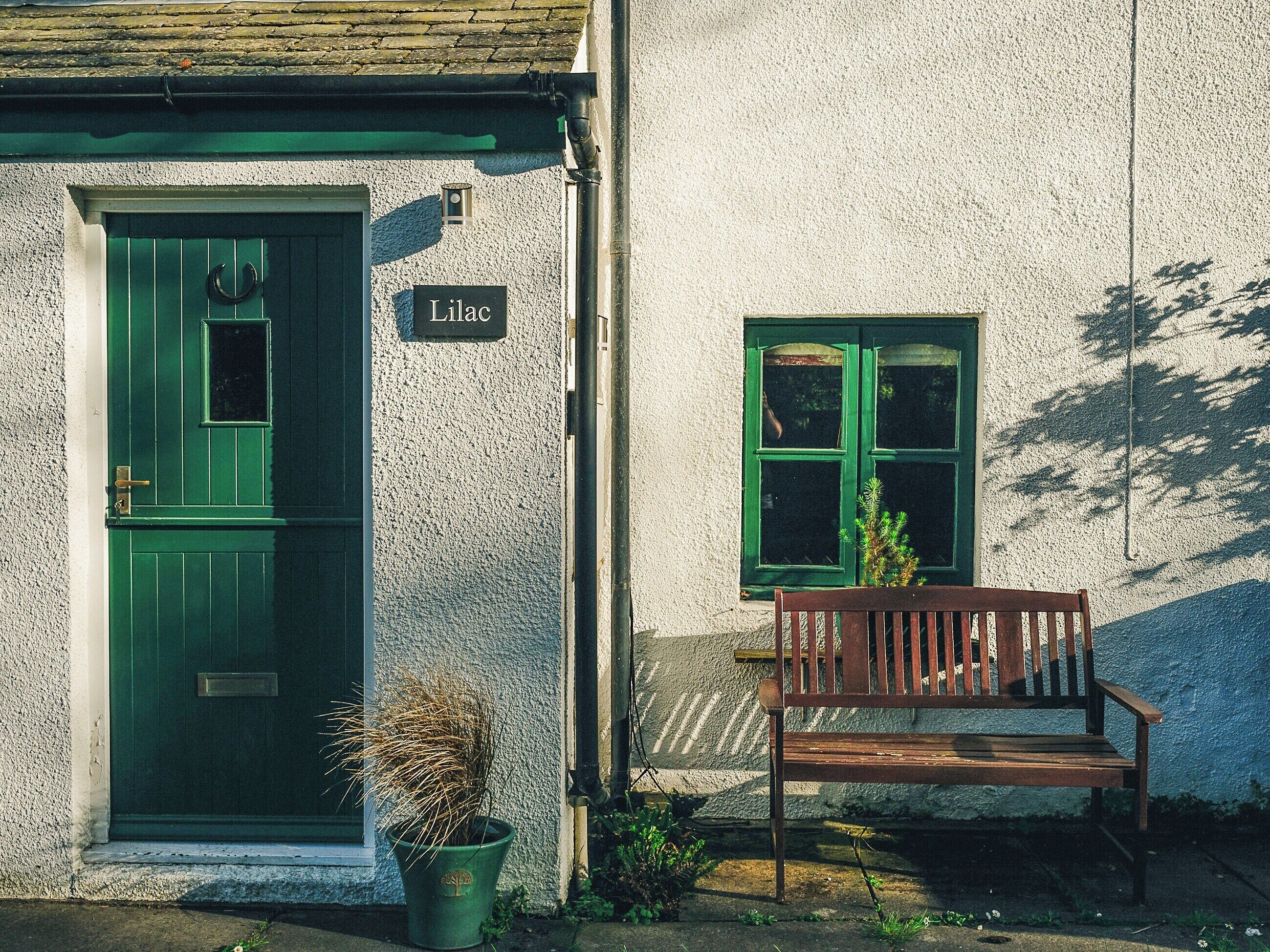 Une porte verte avec une enseigne indiquant « Lilac », un banc et un pot de fleurs devant un bâtiment dont le mur présente une texture particulière.