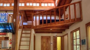Un escalier en bois mène à une chambre aménagée en mezzanine, où se trouvent un lit et une vue sur l’extérieur.