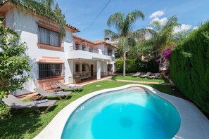 A two-story house with a swimming pool, a patio with lounge chairs, and a well-maintained garden with palm trees and shrubs.