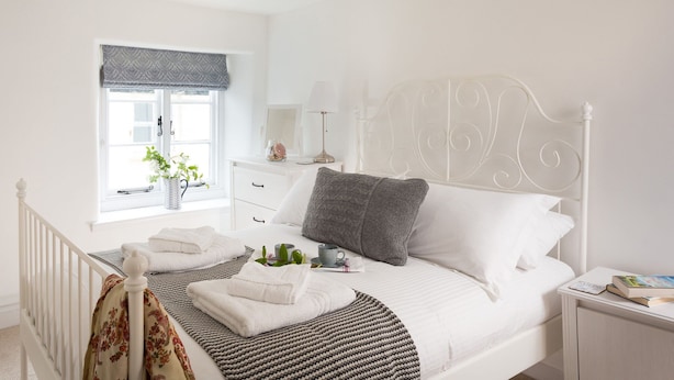 Un lit bien fait, avec du linge de lit blanc, un oreiller gris et une table de chevet sur laquelle se trouvent une lampe et des livres.