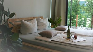 Una cama bien hecha con almohadas, una botella y un vaso sobre una bandeja, con vistas a la vegetación exterior.