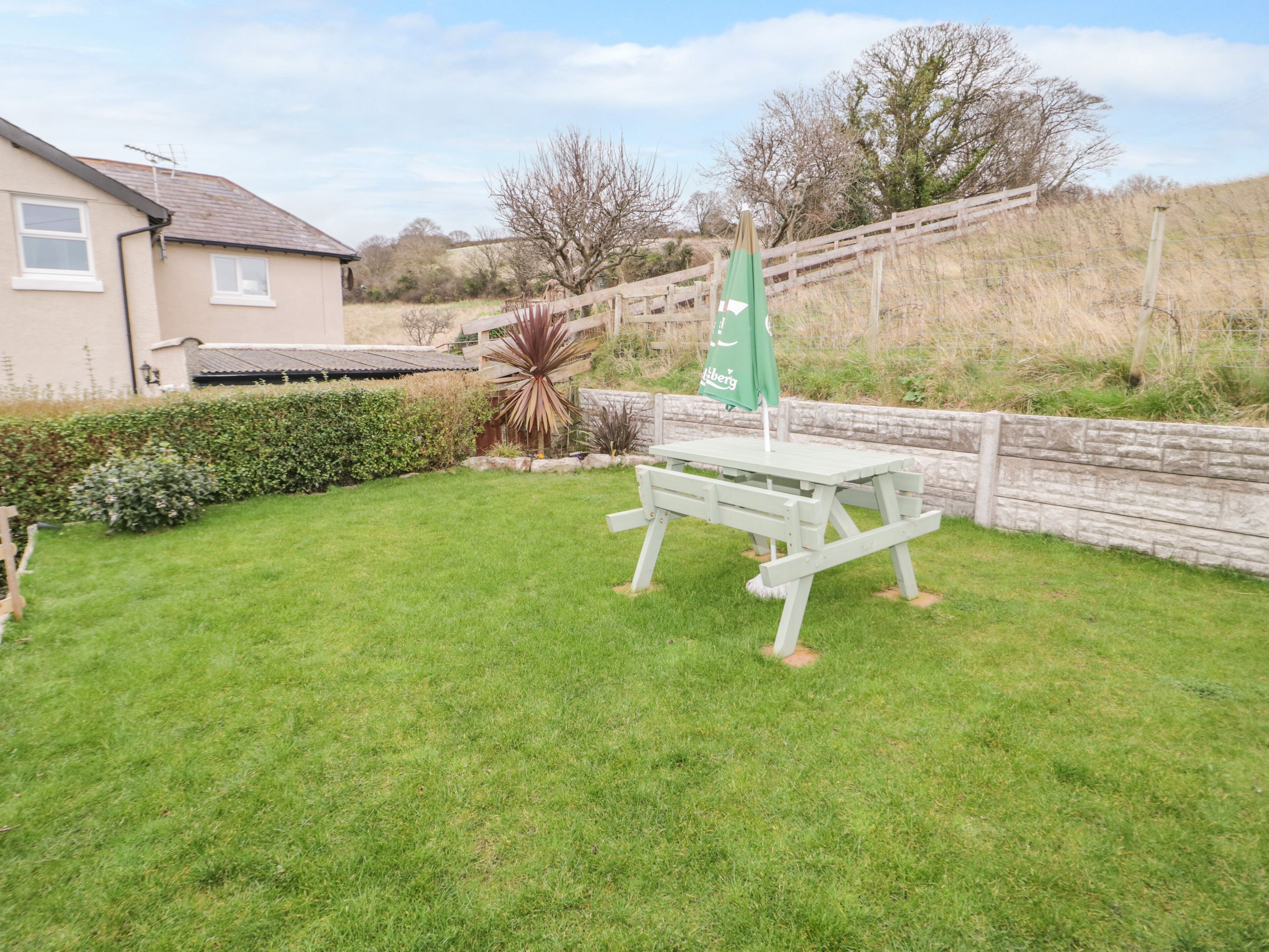 A green picnic table with a green umbrella in a grassy backyard with a stone wall and a wooden fence.