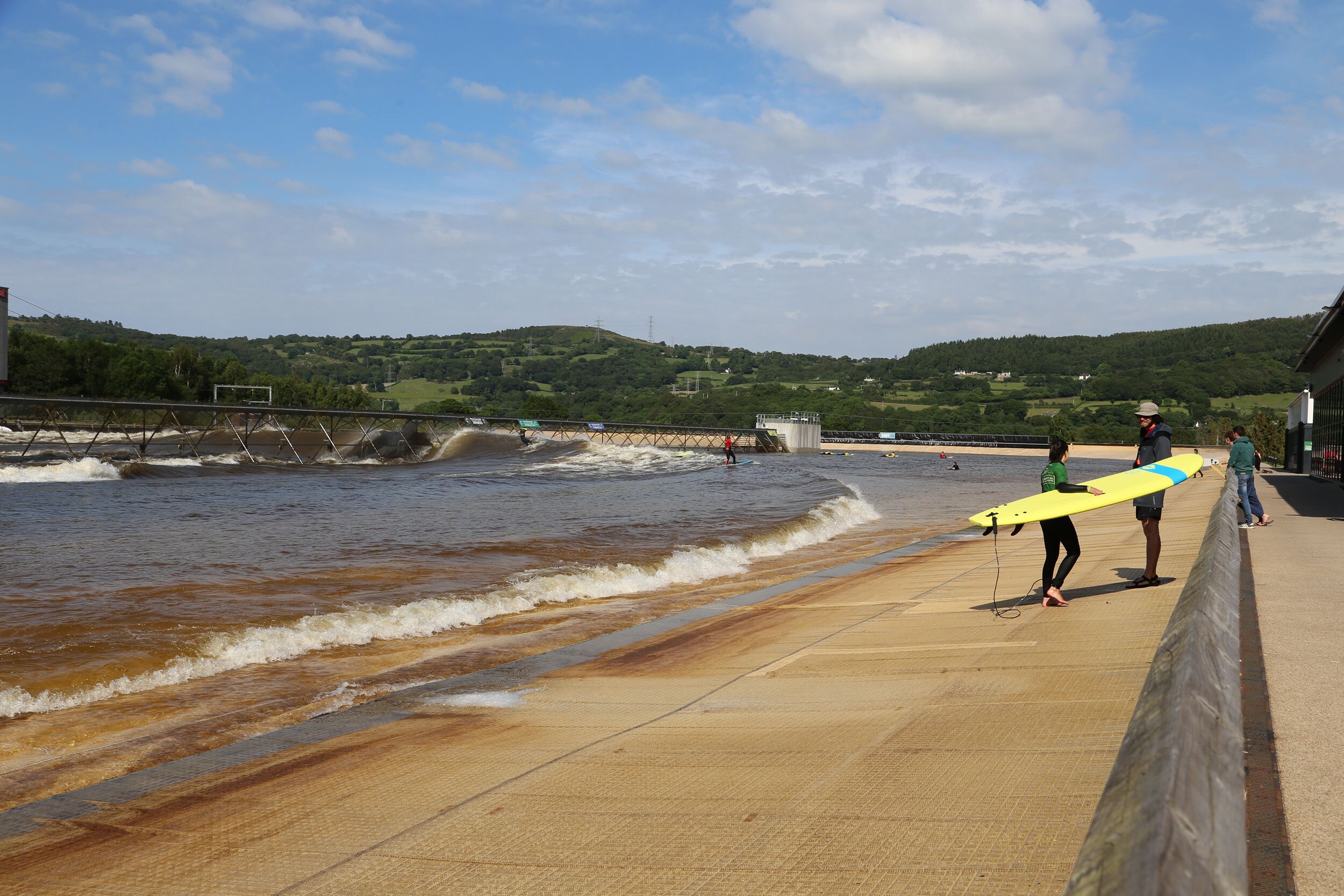 A coastal walkway with a concrete barrier, a beach, and a hilly landscape in the background.