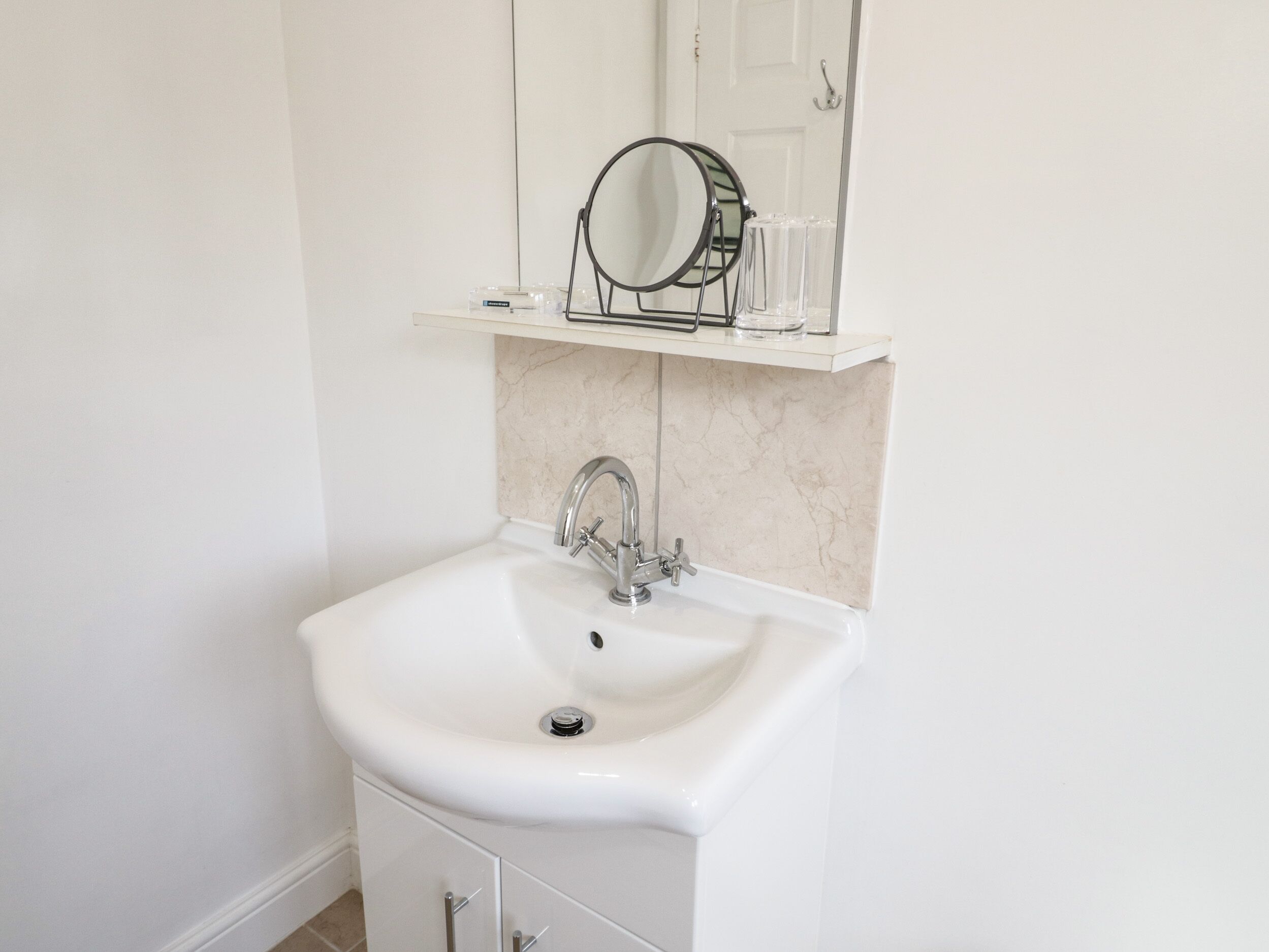A white bathroom with a sink, mirror, and a shelf above the sink.